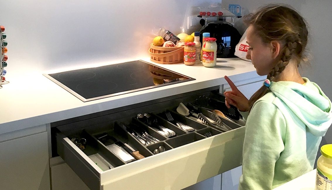 Une fille en sweat vert ouvre un tiroir de cuisine rempli de couverts près d'une plaque de cuisson noire.