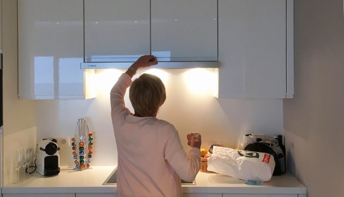 Une personne ajuste une hotte de cuisine sous des placards blancs, avec une machine à café et des capsules colorées à gauche.
