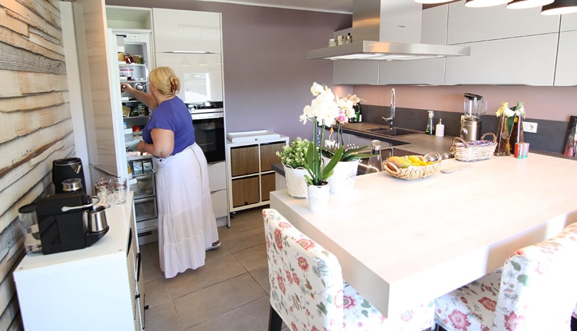Une femme en jupe blanche et t-shirt bleu cherche dans un réfrigérateur ouvert, dans une cuisine moderne avec un îlot central.