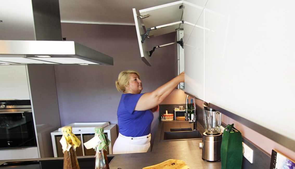 Une femme en haut bleu ouvre un placard blanc dans une cuisine moderne avec un plan de travail gris et un mixeur.