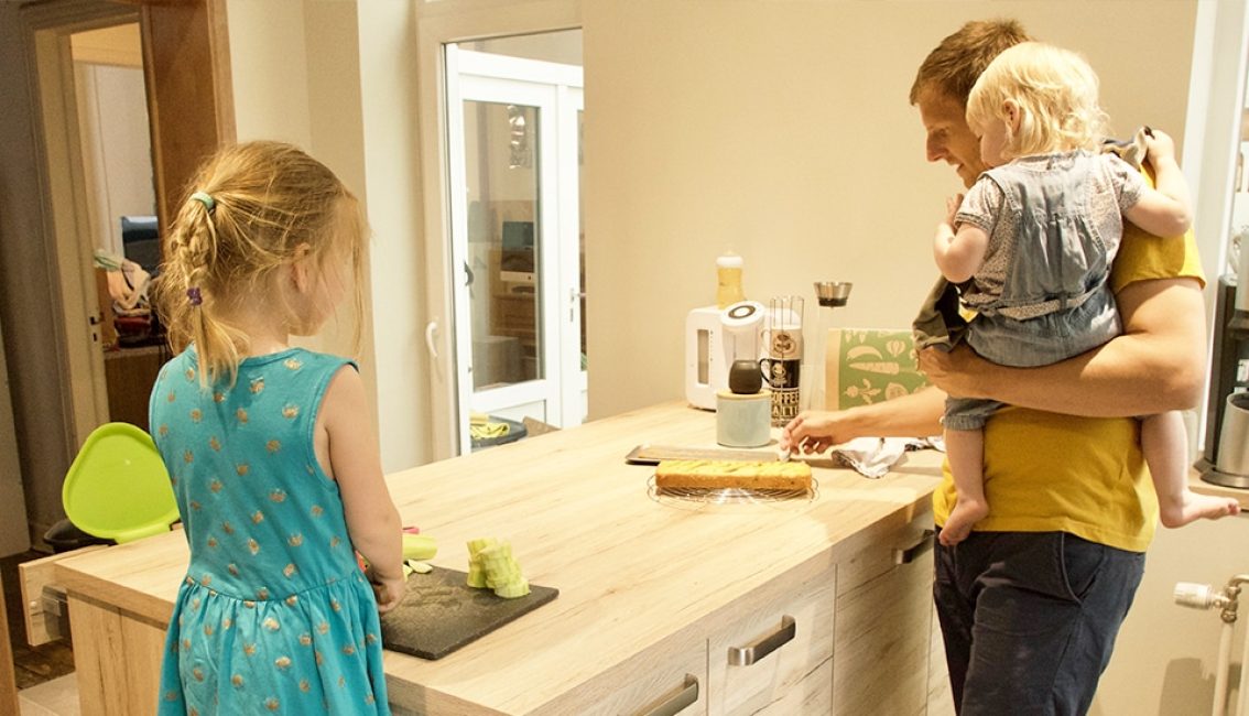 Un homme en t-shirt jaune tient un enfant blond dans une cuisine, tandis qu'une fille en robe bleue se trouve près d'un îlot central en bois clair.