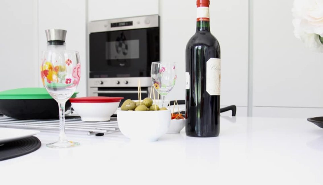 Bouteille de vin rouge, verres à motifs floraux, et bol d'olives sur une table blanche, avec four encastré en arrière-plan.