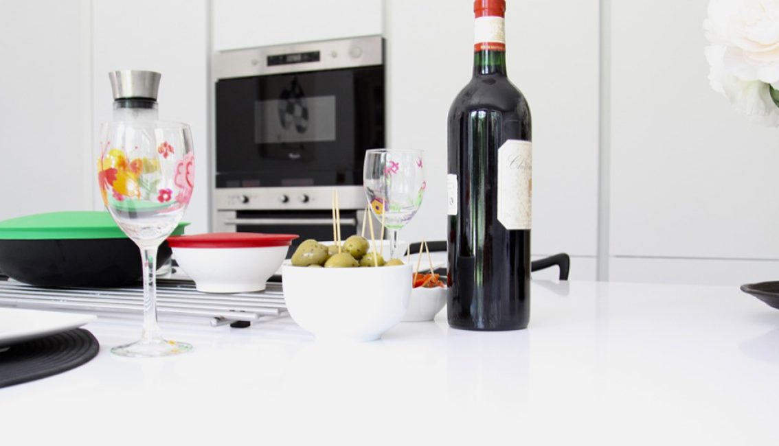 Bouteille de vin rouge, verres à motifs floraux, et bol d'olives sur une table blanche, avec four encastré en arrière-plan.