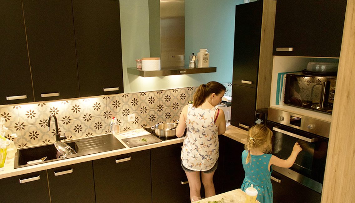 Deux personnes cuisinent dans une cuisine moderne avec des armoires noires, un plan de travail clair et un dosseret à motifs floraux.