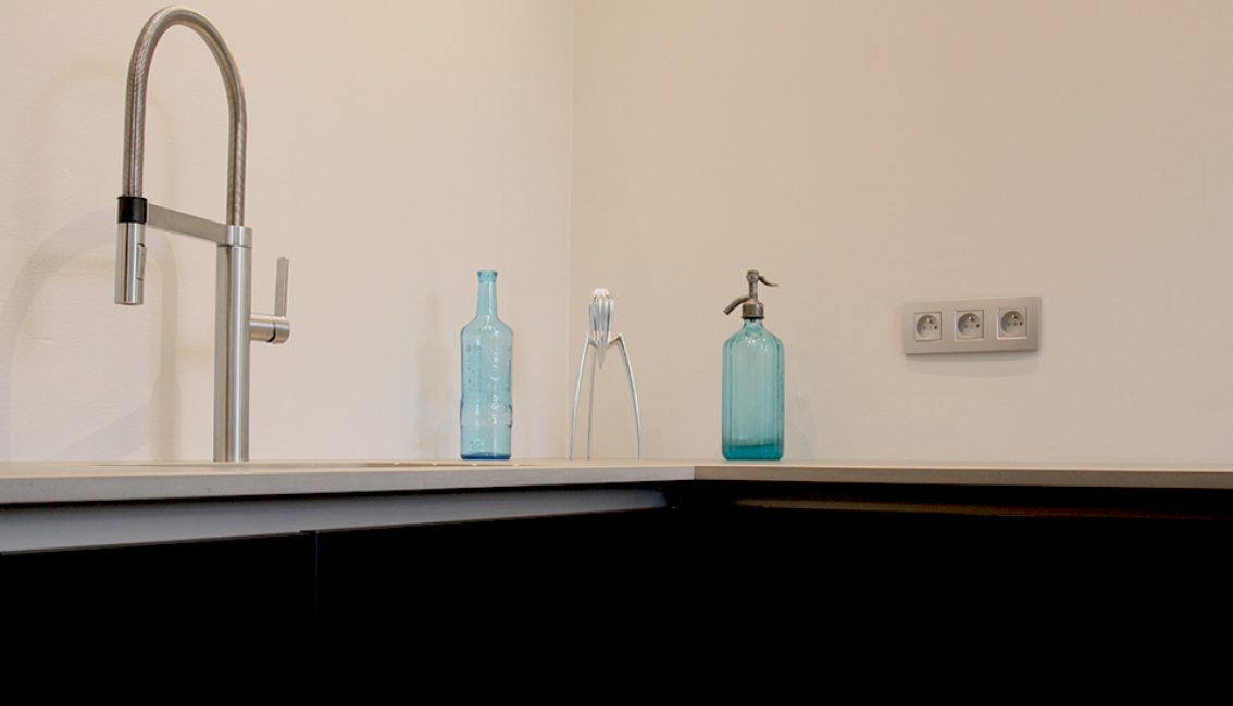 Un robinet en métal, deux bouteilles bleues et un presse-agrumes argenté sont disposés sur un plan de travail beige avec des prises électriques murales.