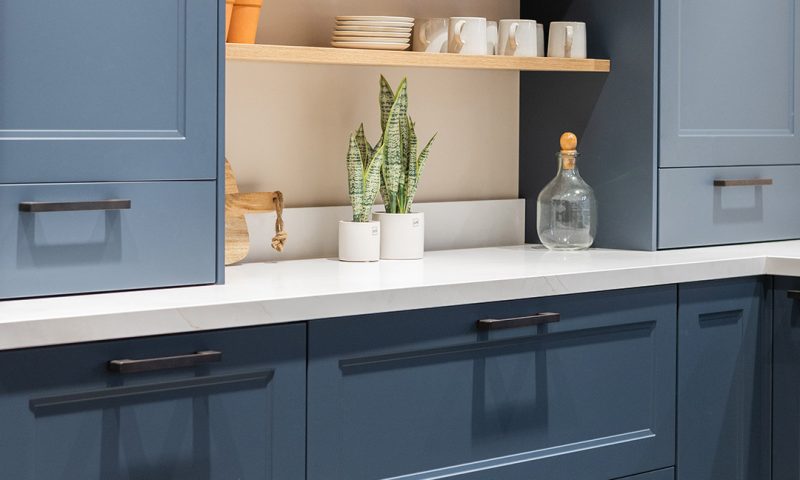Cuisine avec placards bleus et plan de travail blanc, encadrant des étagères en bois clair avec assiettes, tasses blanches, deux plantes en pot et une bouteille en verre.