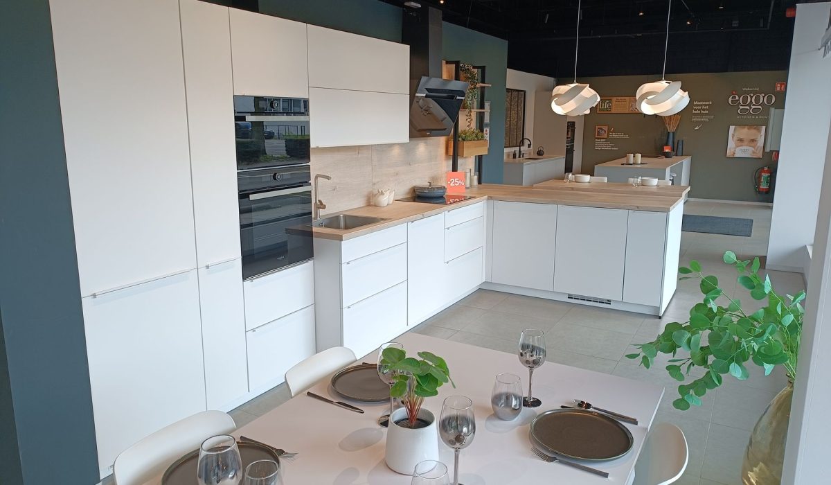 Cuisine d’exposition blanche en L avec plan de travail en bois clair, four encastré noir, évier, table dressée au premier plan et suspensions blanches.