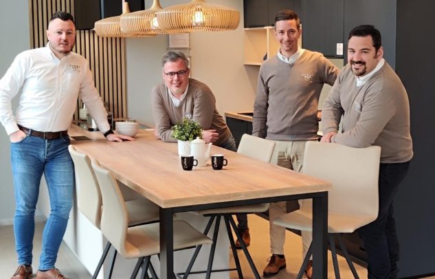 Quatre personnes posent autour d'une table en bois avec des chaises beiges, des tasses noires et une plante verte.