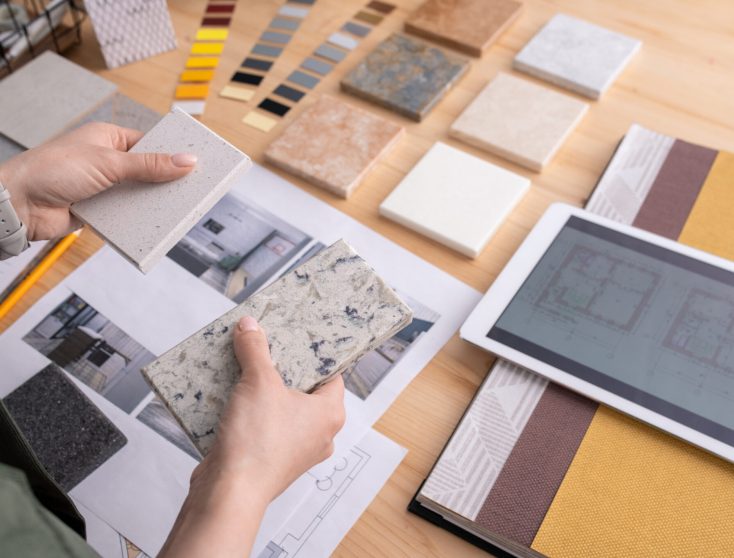 Hands of young female designer holding two samples of marble tile over wooden table with digital tablet, photos of home interior etc