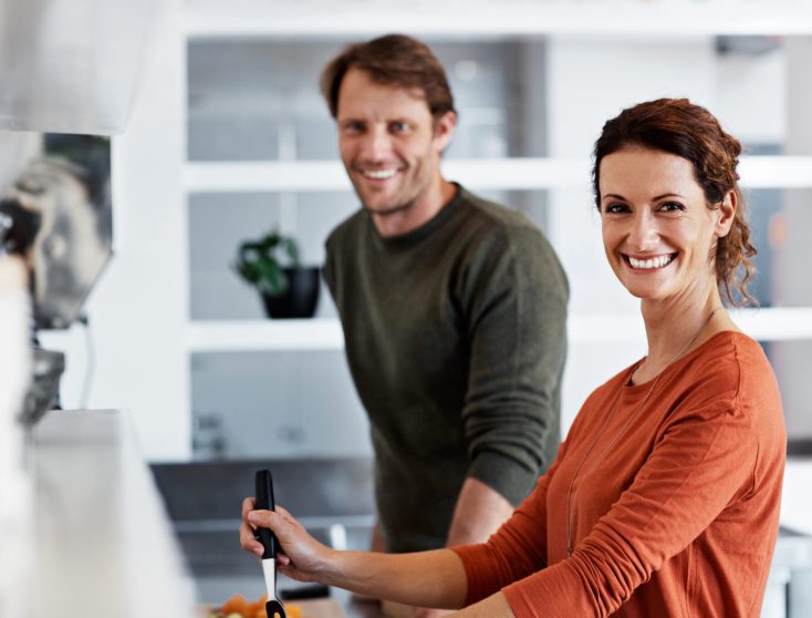 Couple qui sourit dans une cuisine, pendant qu'ils cuisinent ensemble.